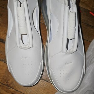 May include: Pair of white athletic shoes with a minimalist design. The shoes feature a smooth, leather-like upper with a subtle Nike logo and a unique lace cover. The soles are white with a visible air pocket.