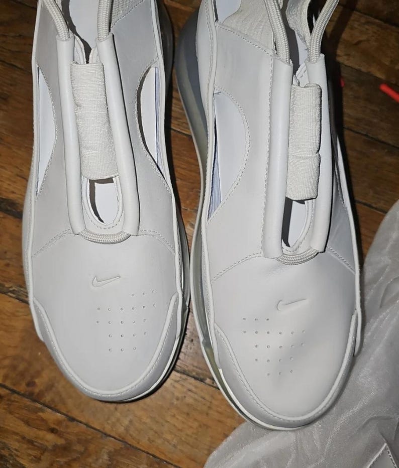May include: Pair of white athletic shoes with a minimalist design. The shoes feature a smooth, leather-like upper with a subtle Nike logo and a unique lace cover. The soles are white with a visible air pocket.