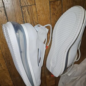 May include: White trainers with a visible air bubble in the sole. The shoes have a strap across the top and a textured sole. The laces are white with a small orange accent. The shoes are on a wooden surface.