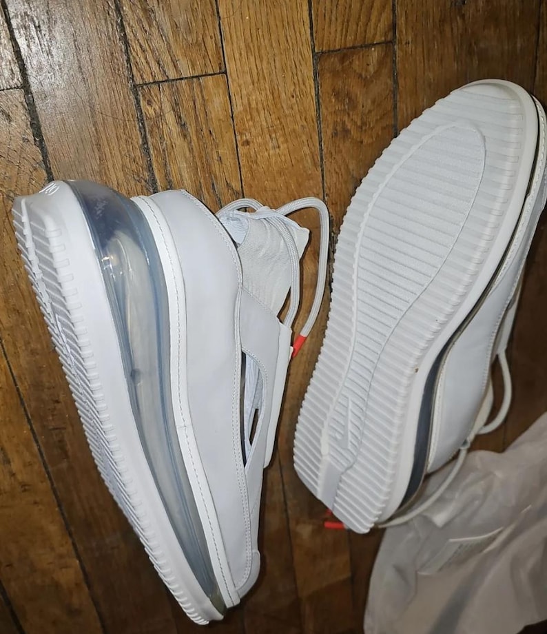 May include: White trainers with a visible air bubble in the sole. The shoes have a strap across the top and a textured sole. The laces are white with a small orange accent. The shoes are on a wooden surface.