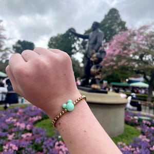 May include: A gold beaded bracelet with a light green Mickey Mouse charm. The bracelet is on a person's wrist.