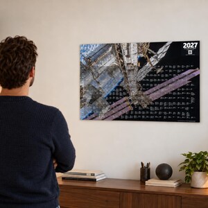 K&ouml;nnte beinhalten: Ein rechteckiger Wandkalender f&uuml;r das Jahr 2027 mit einer hochaufl&ouml;senden Fotografie der Internationalen Raumstation im Erdorbit. Das Design umfasst einen schwarzen Hintergrund mit wei&szlig;em Text und einem QR-Code in der oberen Ecke.