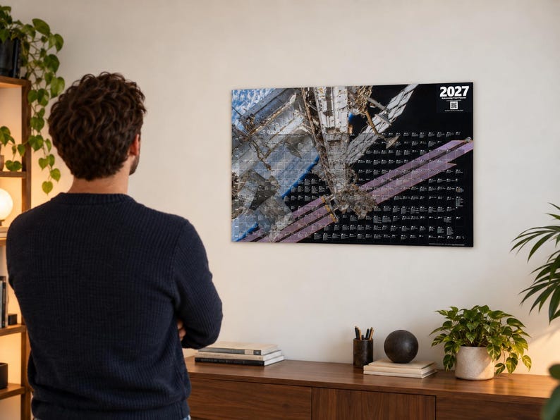 K&ouml;nnte beinhalten: Ein rechteckiger Wandkalender f&uuml;r das Jahr 2027 mit einer hochaufl&ouml;senden Fotografie der Internationalen Raumstation im Erdorbit. Das Design umfasst einen schwarzen Hintergrund mit wei&szlig;em Text und einem QR-Code in der oberen Ecke.
