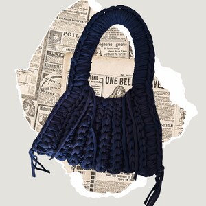 May include: A navy blue, handmade tote bag with a unique, textured design. The bag features a large, looped handle and a body constructed from thick, woven fabric. Tassels hang from the bottom. The background includes torn pieces of vintage newspaper.