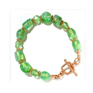May include: A green glass bead bracelet with a copper clasp. The beads are oval shaped and have a light green color with a subtle swirl pattern.