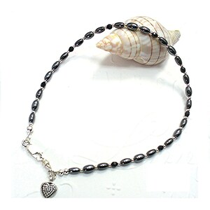 May include: A necklace featuring dark gray oval beads and small black beads. A silver-toned heart pendant with a textured surface hangs from the clasp. The necklace is displayed near a seashell on a white surface.