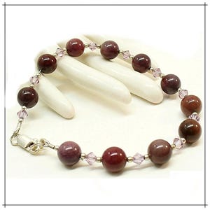 May include: A beaded bracelet featuring large, round, dark red beads interspersed with smaller, light purple crystal beads. The bracelet has a silver clasp and is displayed on a white surface.