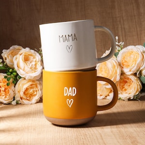 Puede incluir: Dos tazas de cerámica apiladas. La taza superior es blanca con "MAMA" y un corazón. La taza inferior es de color amarillo mostaza con "DAD" y un corazón. Las tazas tienen una base de color neutro y están sobre un fondo de flores.