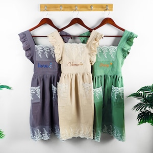 Puede incluir: Tres delantales en tonos grises, beige y verdes cuelgan de perchas de madera. Cada delantal tiene un escote cuadrado, mangas con volantes y un bolsillo. Los delantales están bordados con nombres y diseños florales. Los delantales están ribeteados con encaje blanco.