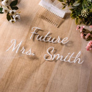 以下が含まれることがあります： 白い筆記体で「Future Mrs. Smith」と刺繍されたブライダルベール。 ベールは櫛に付けられており、花のアクセントに囲まれています。