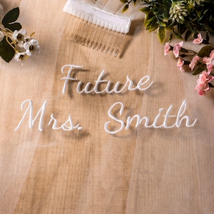 以下が含まれることがあります： 白い筆記体で「Future Mrs. Smith」と刺繍されたブライダルベール。 ベールは透明な櫛に取り付けられており、ニュートラルな背景に配置されています。 花柄のアクセントが見えます。