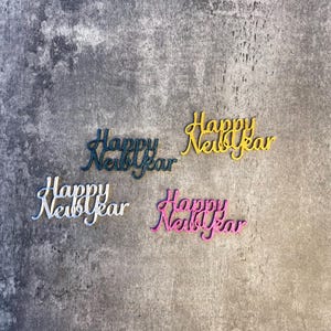 Peut inclure: Quatre découpes cursives "Happy New Year" en teal, jaune, blanc et rose. Les mots sont disposés sur une surface grise texturée. Les découpes sont probablement destinées aux décorations ou aux travaux manuels des fêtes.