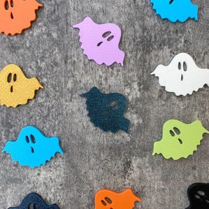 Könnte beinhalten: Eine Ansammlung farbenfroher Geister-Ausschnitte in verschiedenen Farben, darunter Orange, Blau, Grün und Lila. Jeder Geist hat zwei ovale Augenausschnitte und einen wellenförmigen unteren Rand. Die Ausschnitte sind auf einer grau strukturierten Oberfläche verteilt.