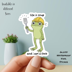 Op de afbeelding: Glanzende, waterdichte vinyl sticker met een cartoon kikker met een gele helm en een vork. De tekst luidt "life is soup and I am a fork". Verkrijgbaar in verschillende maten.
