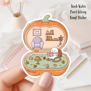 May include: A 3-inch water-proof, glossy vinyl sticker. The sticker features a pumpkin-shaped design with a cozy reading scene inside. The scene includes a character reading a book, a rocking chair, a cozy armchair, and the text "Cozy Reader".