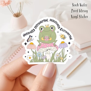 May include: A 3 inch water-proof glossy vinyl sticker featuring a green frog wearing a crown, sitting on a pink and white mushroom with a cup of tea. The frog is surrounded by flowers and butterflies. The sticker reads "Naturally Introverted, Selectively Extroverted".