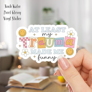 Puede incluir: Una pegatina de vinilo brillante impermeable de 7,6 cm con un diseño colorido que dice "Al menos mi trauma me hizo divertido". La pegatina presenta un patrón a cuadros, caras sonrientes y flores.