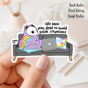 May include: A glossy vinyl sticker featuring a cartoon opossum wrapped in a rainbow blanket, lounging on a gray couch with a laptop. The sticker reads "life hack: play dead to avoid social situations." The sticker is 3 inches in size.