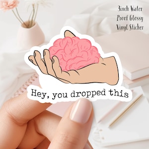 May include: A 3 inch water-proof glossy vinyl sticker with a pink brain in a hand and the text "Hey, you dropped this".