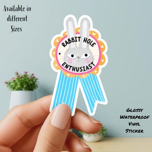 May include: A glossy, waterproof vinyl sticker featuring a cartoon rabbit face with the text "Rabbit Hole Enthusiast." The sticker has a pink and orange scalloped border, blue ribbon, and the text "Available in different Sizes."