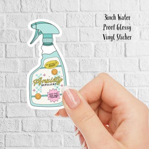 Op de afbeelding: Een 7,6 cm waterdichte, glanzende vinyl sticker van een blauwe en groene spuitfles met de tekst "Anxiety Repellent" en "100% Satisfaction Guaranteed" op het etiket. Het etiket heeft ook een roze en wit geruit patroon en gele smiley's.