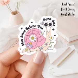 Puede incluir: Una pegatina de vinilo brillante impermeable de 7,6 cm que presenta un caracol rosa con un símbolo de paz en su caparazón. El caracol está rodeado de flores y hongos. El texto "Slow Down Before You Burn Out" está escrito alrededor del caracol.