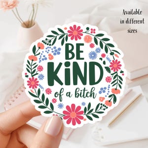 May include: A round sticker with a floral wreath design. The sticker features the words "BE KIND of a bitch" in dark green letters. The wreath includes pink, blue, and orange flowers with green leaves. Text on the right says "Available in different sizes."