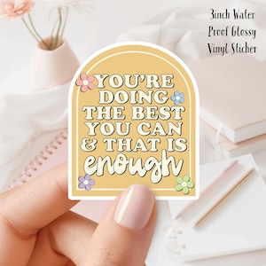 May include: A 3 inch waterproof glossy vinyl sticker with a yellow background and the text "YOU'RE DOING THE BEST YOU CAN & THAT IS enough" in a retro font. The sticker is decorated with colourful flowers.