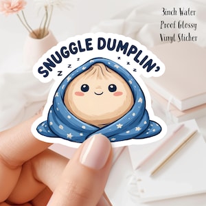 May include: A 3-inch water-proof glossy vinyl sticker featuring a cartoon dumpling wrapped in a blue blanket with white stars. The sticker has the text "Snuggle Dumplin'" in white, with "z" symbols above the dumpling.