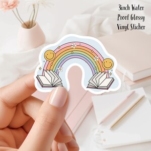 May include: A 3 inch water-proof glossy vinyl sticker featuring a rainbow with two open books and smiley faces on either side. The rainbow is in shades of pink, orange, yellow, green, blue, and purple.