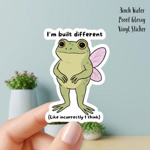 May include: A 3-inch water-proof glossy vinyl sticker featuring a cartoon frog with fairy wings. The frog is light green with large eyes and the text "I'm built different (Like incorrectly I think)".