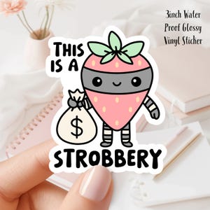 May include: A glossy vinyl sticker featuring a cartoon strawberry wearing a black mask and striped pants, holding a money bag with a dollar sign. The text reads "THIS IS A STROBBERY". The sticker is 3 inches in size.