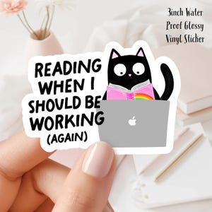 May include: A black cat wearing a pink shirt sits on a laptop computer and reads a book. The text on the sticker reads "Reading when I should be working (again)".