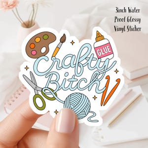 May include: A 3-inch water-proof, glossy vinyl sticker. The sticker features the words "Crafty Bitch" in a whimsical font, surrounded by craft-related illustrations like a paint palette, scissors, yarn, and glue.