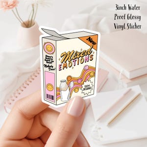 Puede incluir: Una pegatina de vinilo brillante impermeable de 7,6 cm que representa una caja de cereales con el texto "Mixed Emotions" y una cara sonriente de dibujos animados. La caja es rosa y naranja con fondo blanco. El texto "New" está en una explosión de estrellas rosa. El texto "100% Organic" está en un círculo con un borde rosa y naranja.