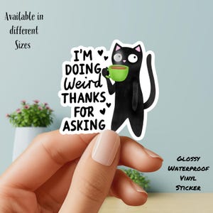 May include: A glossy, waterproof vinyl sticker featuring a black cat holding a green mug. The cat has one large eye and the text "I'M DOING Weird THANKS FOR ASKING" is displayed. The sticker is available in different sizes.
