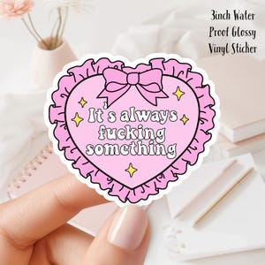 May include: A pink heart-shaped vinyl sticker with the phrase "It's always fucking something" in white text. The sticker has a ruffled border, a bow, and four yellow star accents. The sticker is 3 inches in size.