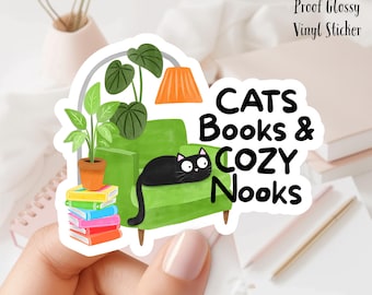 Könnte beinhalten: Ein glänzender Vinyl-Aufkleber mit einer schwarzen Katze auf einem grünen Sessel, mit dem Text "CATS Books & COZY Nooks". Der Aufkleber enthält auch Illustrationen von Pflanzen, Büchern und einer Lampe. Der Aufkleber ist ca. 7,6 cm groß.