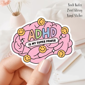 Puede incluir: Una pegatina de vinilo rosa con forma de cerebro con la palabra "ADHD" en letras coloridas. El texto "IS MY SUPER POWER" está debajo de la palabra ADHD. La pegatina tiene caras sonrientes amarillas y acentos de estrellas. La pegatina mide 7,6 cm.