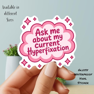 May include: A pink and white vinyl sticker with the text "Ask me about my current Hyperfixation" in a bubble design. The sticker is surrounded by pink diamond shapes and the words "Glossy Waterproof Vinyl Sticker".