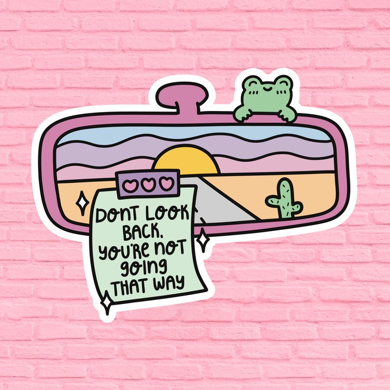 May include: A pink and white sticker with a cartoon rearview mirror showing a sunset over a desert road. A green frog sits on top of the mirror. A green note attached to the mirror says "Don't look back. You're not going that way."