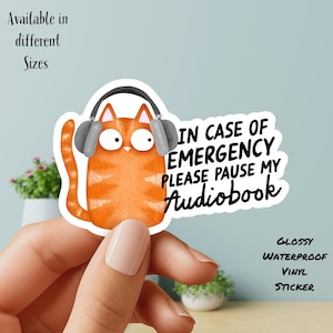 May include: A glossy, waterproof vinyl sticker featuring an orange cartoon cat wearing headphones. The sticker reads "IN CASE OF EMERGENCY PLEASE PAUSE MY Audiobook." Additional text states "Available in different Sizes" and "GLOSSY WATERPROOF VINYL STICKER."
