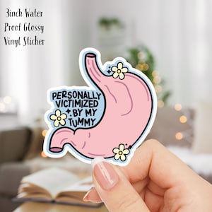 Peut inclure: Un autocollant en vinyle brillant imperméable de 7,6 cm avec un estomac rose en dessin animé et le texte "PERSONALLY VICTIMIZED BY MY TUMMY" avec des fleurs jaunes.