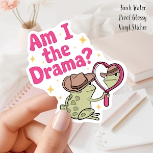 Könnte beinhalten: Ein 7,6 cm großer, wasserfester, glänzender Vinyl-Aufkleber mit einer Cartoon-Frosch, die einen Cowboyhut trägt und sich in einem herzförmigen Spiegel betrachtet. Der Text "Am I the Drama?" ist in rosa Buchstaben über dem Frosch geschrieben.