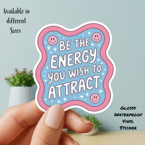 May include: A glossy, waterproof vinyl sticker with a wavy pink border and a light blue interior. The sticker features the text "BE THE ENERGY YOU WISH TO ATTRACT" in white, along with smiley faces and star accents. The text "Available in different Sizes" is also visible.