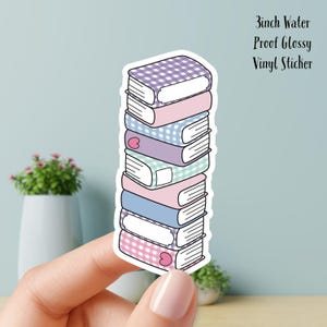 Op de afbeelding: Een sticker met een stapel kleurrijke boeken. Elk boek heeft een andere kleur en patroon, waaronder ruitjes en effen kleuren. De sticker is 7,6 cm groot en gemaakt van waterdicht, glanzend vinyl. De boeken hebben witte labels.