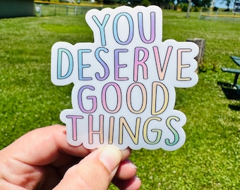 May include: A white sticker with the text "YOU DESERVE GOOD THINGS" in rainbow colours. The sticker is being held in a hand against a green grassy background.