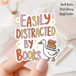 Könnte beinhalten: Ein weißer Vinyl-Aufkleber mit einem rosa, orangefarbenen und gelben Design. Der Aufkleber zeigt eine Cartoon-Gans, die einen Stapel Bücher trägt, und den Text "Easily Distracted by Books".