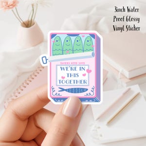 May include: A 3-inch waterproof glossy vinyl sticker with a pink and blue design. The sticker features a cartoon illustration of a can of sardines with the text "Packed with Love" and "We're in this together".