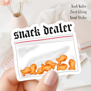 May include: A glossy vinyl sticker featuring a clear plastic bag design with the words "Snack Dealer" in a gothic font. The bag contains orange goldfish crackers. The sticker is 3 inches in size and water proof.